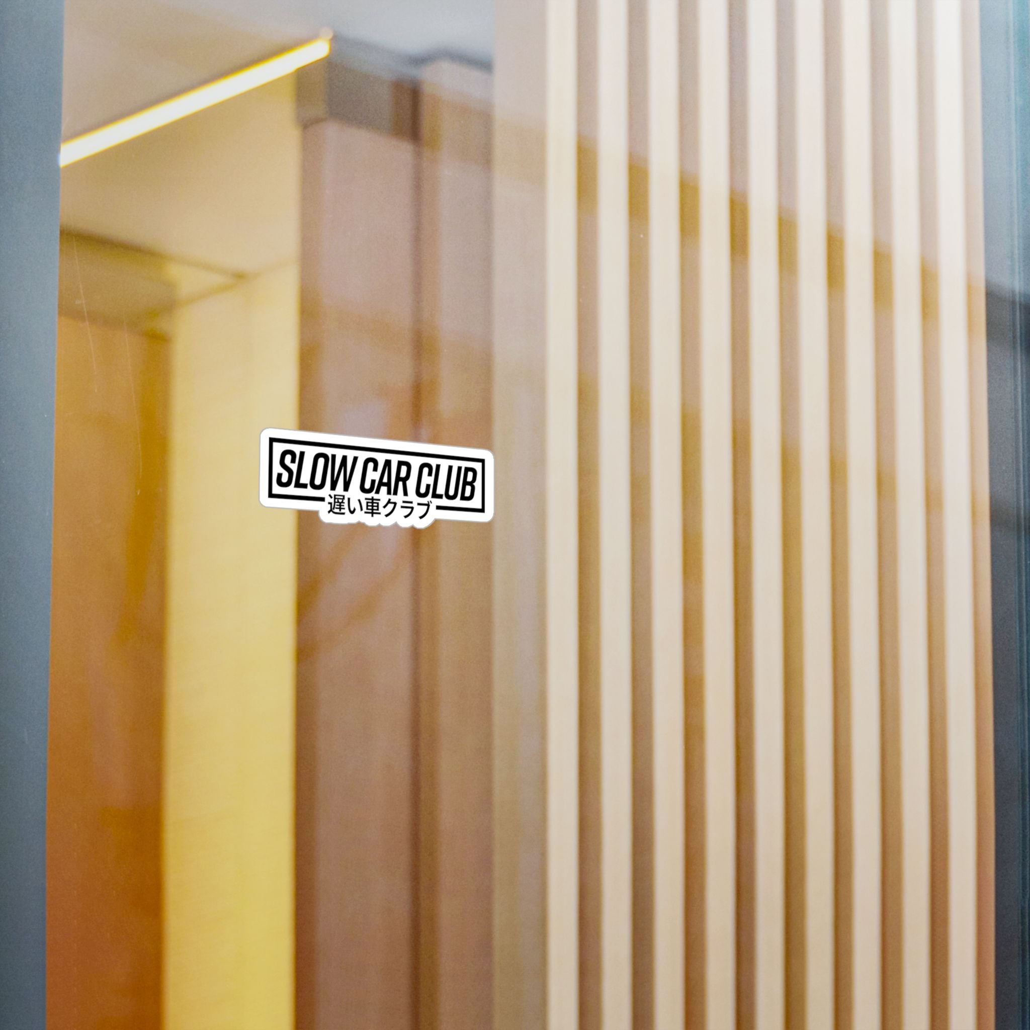 A rectangular sticker with the text 'SLOW CAR CLUB' and Japanese characters below it, shown on a glass door or window with warm reflections and vertical slats in the background.