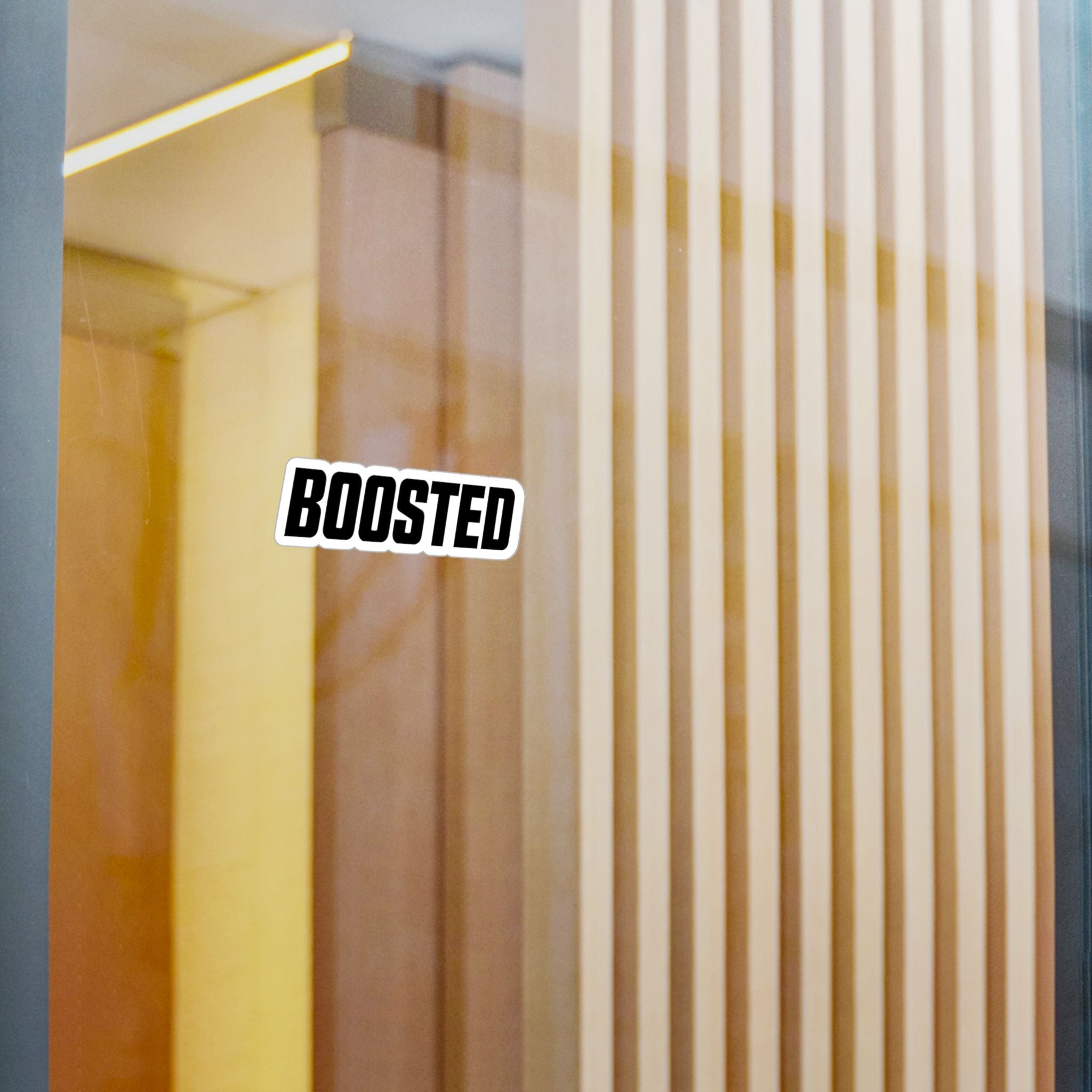 A die-cut vinyl sticker with the word 'BOOSTED' in bold black letters on a white background with a black outline, applied to a glass door or window with a warm, modern interior visible behind it.
