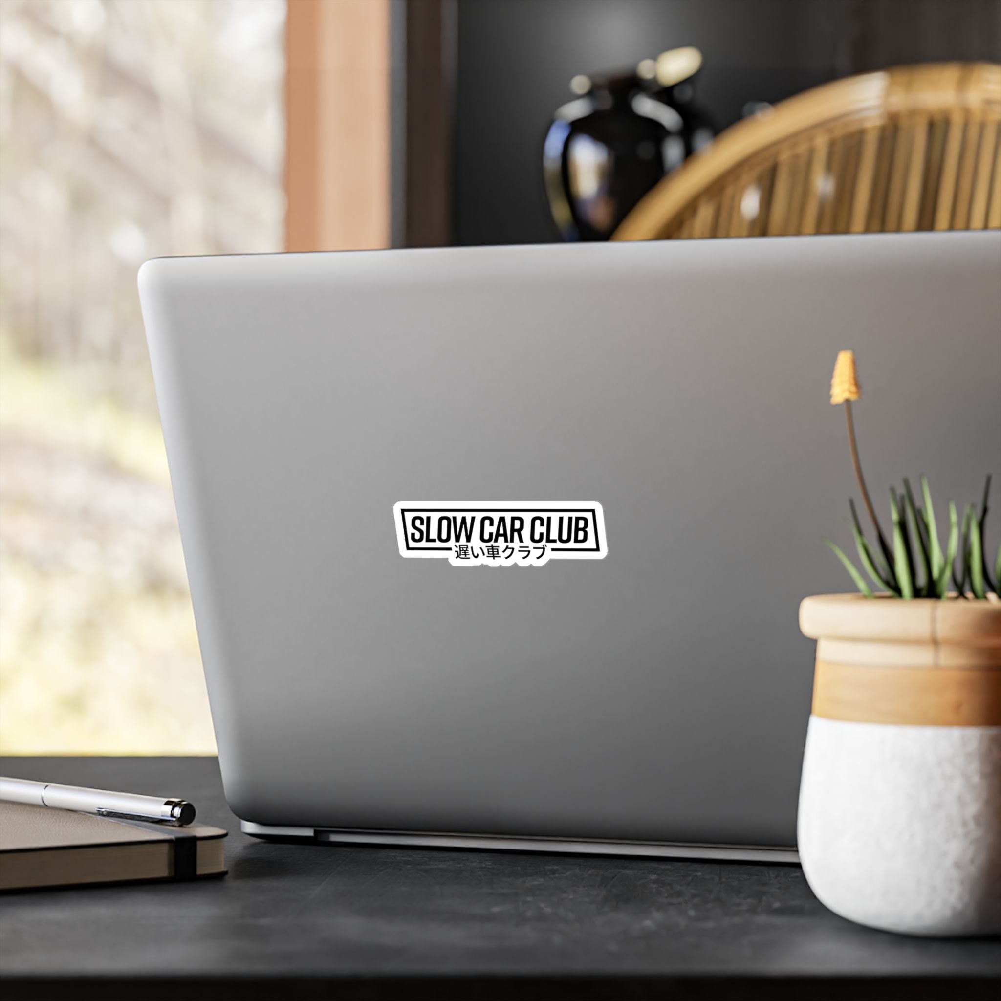 Laptop with a sticker " Slow Car Club" on a desk with a plant and pen